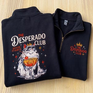 May include: Two navy blue sweatshirts with a zipper and a high collar. One features a cat wearing sunglasses and a crown, holding a cocktail, with the text "THE DESPERADO CLUB SO FUN IT HURTS." The other has the text "THE DESPERADO CLUB" with a crown.