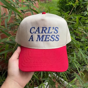 May include: A baseball cap with a cream-colored crown and a red brim. The text "CARL'S A MESS" is embroidered in blue on the front. The cap has a red button on top.