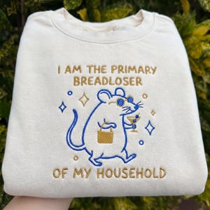 May include: Cream-colored sweatshirt with the embroidered phrase "I AM THE PRIMARY BREADLOSER OF MY HOUSEHOLD." The design features a blue cartoon mouse wearing sunglasses and holding a cocktail and a small bag. Decorative blue stars and diamonds surround the mouse.
