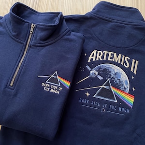 May include: Two navy blue quarter-zip sweatshirts. One features a prism with a rainbow and the text "DARK SIDE OF THE MOON." The other has an image of the moon, a satellite, and the text "ARTEMIS II" and "DARK SIDE OF THE MOON."