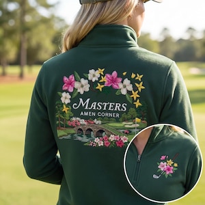 May include: Dark green quarter-zip sweatshirt featuring a floral and golf course design. The back of the sweatshirt displays the words "Masters Amen Corner" with a bridge and flowers. A smaller design of flowers and a golf club is on the chest.