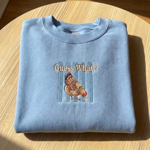 May include: A light blue sweatshirt with the embroidered phrase "Guess What?" above a detailed hen illustration. The hen is brown, tan, and red, with a blue striped background.