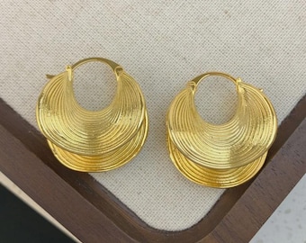 May include: A pair of gold-coloured hoop earrings. Each earring has a layered, fan-like design with a textured surface. The earrings are displayed on a light-coloured fabric inside a wooden tray.