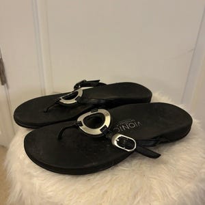 May include: A pair of black Vionic Orthoheel flip-flops with silver-toned metal accents. The sandals have a thong-style design with a buckle closure and are resting on a white, fluffy surface.