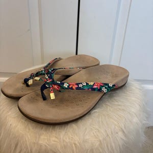 May include: A pair of beige flip-flops with floral straps. The straps are navy blue with a pattern of pink, orange, and white flowers. The flip-flops are resting on a white, fluffy ottoman.