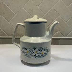 May include: A light blue ceramic teapot with a lid and handle. The teapot features a floral design with white and yellow flowers and blue accents. The teapot is a vintage style and is suitable for serving tea.