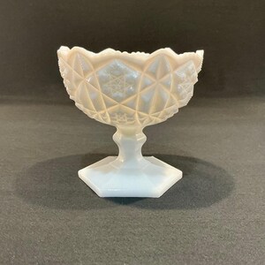 May include: A white, vintage-style glass bowl with a diamond pattern and scalloped rim. The bowl sits on a pedestal base with a hexagonal foot. The glass has a pearlescent sheen, reflecting light.