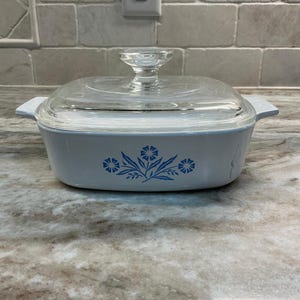 May include: A light blue square casserole dish with a clear glass lid. The dish features a floral design in blue. The lid has a clear glass handle. The dish is sitting on a marble countertop.