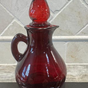 May include: A ruby red glass cruet with a handle and a teardrop-shaped stopper. The cruet has a rounded body and a small spout. The stopper is textured and the cruet is sitting on a black surface.