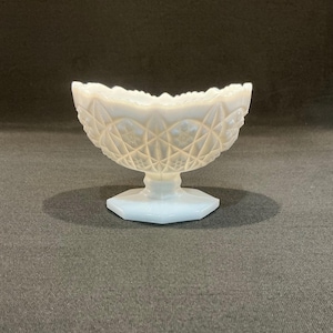 May include: A white, vintage glass dessert dish with a scalloped rim and a geometric pattern. The bowl sits on a hexagonal base and is likely made of milk glass. The dish is likely from the mid-20th century.