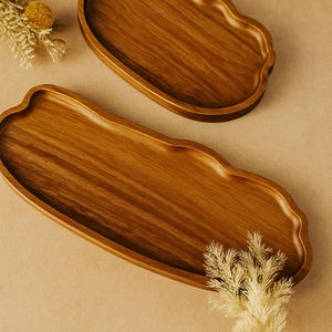 以下が含まれることがあります： 波型の縁が付いた2つの木製サービングトレイ。トレイは明るい茶色の木材でできており、滑らかな仕上がりです。トレイは空で、ニュートラルな背景に置かれています。装飾用の乾燥植物がトレイの近くに配置されています。