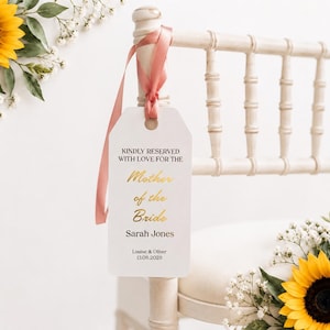 May include: A white rectangular tag with rounded corners hangs from a chair by a dusty rose ribbon. The tag features black text reading KINDLY RESERVED WITH LOVE FOR THE, gold script text reading Mother of the Bride, and black text Sarah Jones, Louise & Oliver, 13.08.2028.