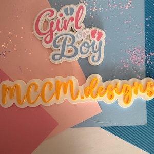 Puede incluir: Un diseño en capas con las palabras "Girl or Boy" en rosa y azul, con un gráfico de huellas. Debajo, el texto "mccm.designs" está en escritura naranja. El fondo presenta papel rosa, blanco y azul con purpurina.