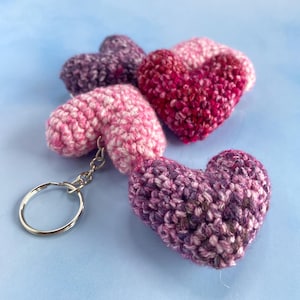 May include: A collection of handmade crochet hearts in shades of pink and purple. One heart is attached to a silver keychain. The hearts are arranged on a light blue surface, showcasing the texture and color variations.