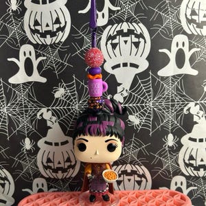 以下が含まれることがあります： ハロウィーンをテーマにしたFunko Popフィギュア。黒と紫の髪の毛のキャラクターがボウルを持っています。フィギュアの上には、ピンクのグリッターボール、"Hocus Pocus"の文字が入った紫色のマグカップ、その他の装飾ビーズが付いたチャームが付いています。背景は、白いハロウィーンをテーマにしたパターンが施された黒色です。