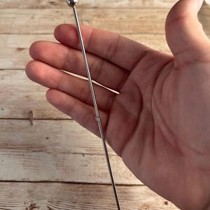 Peut inclure: Un pilon à cocktail en acier inoxydable avec une petite boule ronde au sommet et une extrémité pointue. Le pilon est tenu dans une main sur un fond en bois brun clair. L'outil est conçu pour mélanger les boissons.