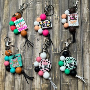 May include: Keychains with various designs. One keychain has the text "Handy Honey" on a white background. Another has a cow print design. Other keychains have the text "How I feel like a teacher" and a mushroom design.