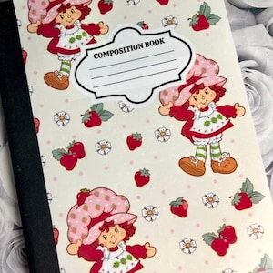 May include: A composition book featuring a repeating pattern of a cartoon character in a strawberry hat and dress, strawberries, and flowers on a cream background. The cover includes a label that reads "COMPOSITION BOOK". The spine is black.