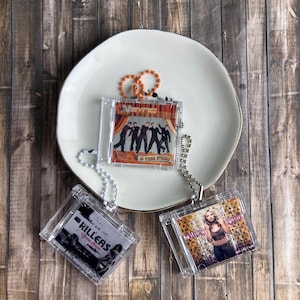 May include: Three clear plastic CD case keychains featuring album art. One keychain displays *NSYNC, another *The Killers*, and the third *Britney Spears*. Each keychain has a beaded chain and a small orange or silver ring.