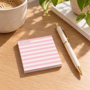 May include: A square notepad featuring a pattern of horizontal light pink and white stripes. The top left corner of the notepad includes the printed text "Just remind me..." in a dark blue script font. The notepad is positioned on a wooden surface.
