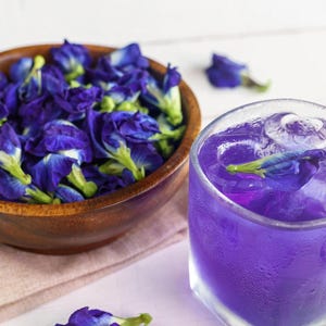 May include: A wooden bowl brimming with vivid blue butterfly pea flowers is placed beside a glass of purple drink with ice. Scattered flowers decorate the white surface, creating a refreshing and visually appealing scene.