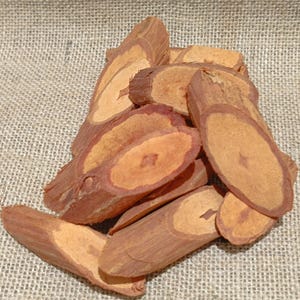 May include: A collection of reddish-brown wood pieces, possibly bark or small logs, with lighter interiors. The wood has a natural, organic appearance, with visible grain and texture. The pieces are arranged on a rough, beige-coloured surface.