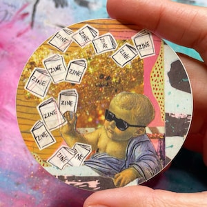 May include: A round sticker featuring a baby wearing sunglasses, surrounded by floating zines. The zines have the word "ZINE" printed on them. The background has gold glitter and colorful abstract patterns.