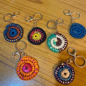 May include: A collection of handmade keychains with circular woven designs in various colors. Each keychain features a gold-colored clasp and ring. The keychains display a range of colors, including blue, orange, black, green, and purple, with intricate patterns.