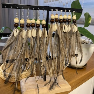 May include: Handmade earrings with a bohemian design. Each earring combines natural elements: wooden beads, seashells, and brown feathers. The earrings are displayed on a black metal stand, highlighting their intricate design and earthy tones.