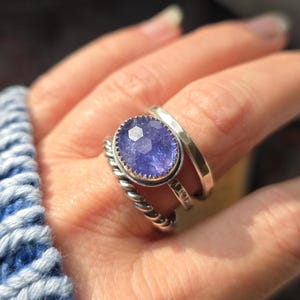 May include: A stack of three silver rings featuring a central oval-shaped faceted purple gemstone in a serrated bezel setting. The stack includes one ring with a twisted rope texture, one plain band, and one ring with a hammered finish surface.