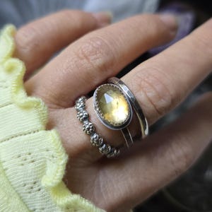 May include: A hand displays three silver rings. The largest ring features an oval, yellow gemstone set in a silver bezel. A hammered silver band and a beaded ring complete the set. The rings are worn on a finger.