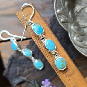 May include: A pair of silver drop earrings, each featuring three oval turquoise stones set in metal bezels. The earrings are displayed against a wooden ruler, showing a length of approximately 2 inches from the top hook to the bottom stone.