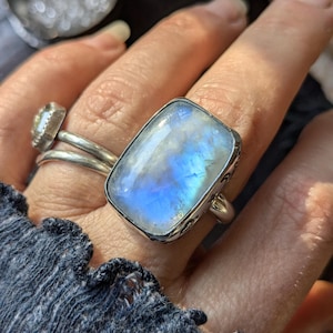 May include: A silver ring with a large, rectangular, iridescent moonstone. The stone has a blue and white color.