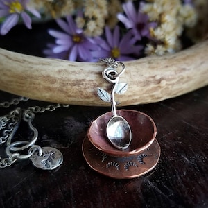 May include: A silver spoon necklace with a rose design and a copper cup pendant. The cup has a textured surface and a crescent moon design.