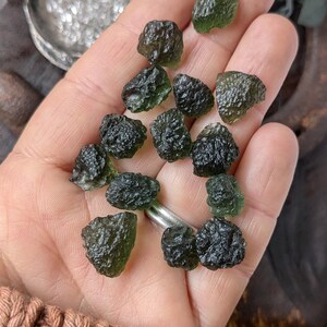 May include: A collection of dark green and black tektite stones, also known as Moldavite, in a hand. The stones are rough and have a glassy texture.