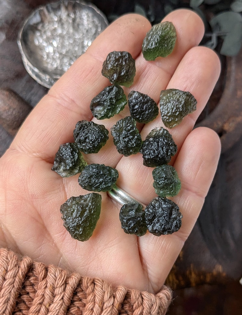 May include: A collection of dark green and black tektite stones, also known as Moldavite, in a hand. The stones are rough and have a glassy texture.