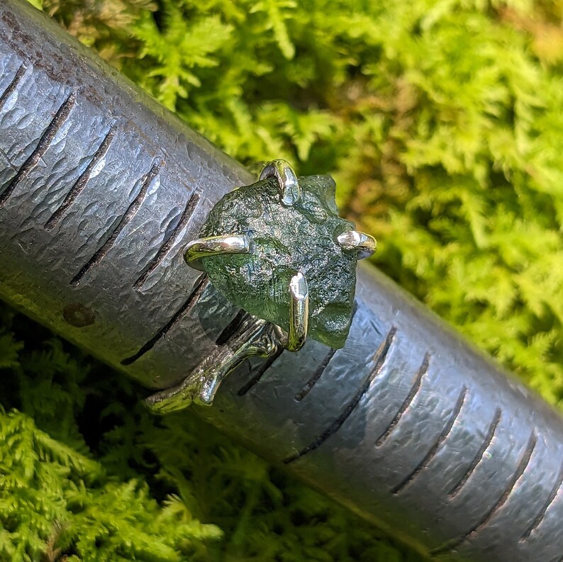 May include: A silver ring with a green, rough, gemstone set in a four-prong setting. The ring is on a metal surface.