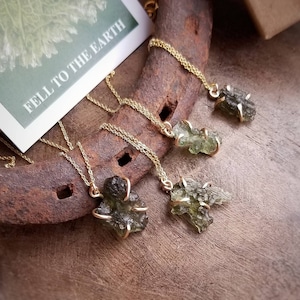 May include: Four gold necklaces with green gemstone pendants. The necklaces are displayed on a rustic wooden surface. The text "Fell to the Earth" is visible on a card in the background.