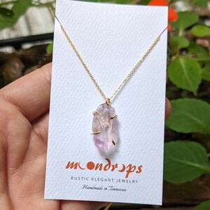 Pu&ograve; includere: Una delicata collana a catena dorata con un ciondolo in cristallo di ametista grezzo. Il cristallo &egrave; di colore viola chiaro e ha una texture naturale e ruvida. La collana &egrave; su una carta bianca con il testo "moondrops rustic elegant jewelry handmade in Tennessee".
