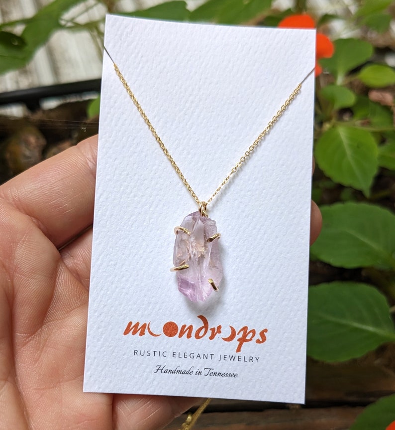 Pu&ograve; includere: Una delicata collana a catena dorata con un ciondolo in cristallo di ametista grezzo. Il cristallo &egrave; di colore viola chiaro e ha una texture naturale e ruvida. La collana &egrave; su una carta bianca con il testo "moondrops rustic elegant jewelry handmade in Tennessee".