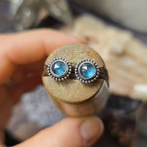 Peut inclure: Une paire de boucles d'oreilles à clous en argent avec des pierres bleues rondes et facettées. Chaque boucle d'oreille est entourée d'une bordure argentée perlée. Les boucles d'oreilles sont présentées sur un objet cylindrique brun.