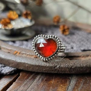Peut inclure: Bague en argent ornée d'une pierre rouge en forme de cœur, facettée. La pierre est entourée d'une bordure argentée perlée. La bague est posée sur une surface texturée, avec un arrière-plan flou.