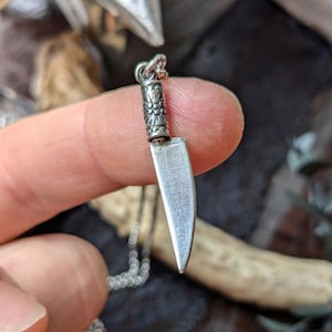 May include: A silver-colored pendant necklace featuring a miniature cleaver charm. The charm has a detailed handle and a small, sharp blade. The necklace chain is also silver. The pendant is held by a finger.