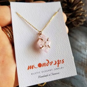 Pu&ograve; includere: Una delicata collana a catena dorata con un ciondolo di cristallo rosa. Il cristallo &egrave; avvolto in filo d'oro e ha una texture naturale e ruvida. La collana &egrave; su una carta bianca con il testo "moondrops rustic elegant jewelry handmade in tennessee".