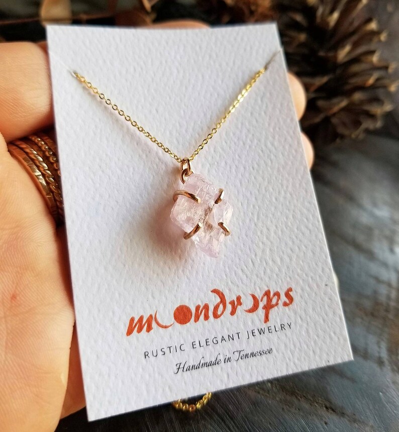 Pu&ograve; includere: Una delicata collana a catena dorata con un ciondolo di cristallo rosa. Il cristallo &egrave; avvolto in filo d'oro e ha una texture naturale e ruvida. La collana &egrave; su una carta bianca con il testo "moondrops rustic elegant jewelry handmade in tennessee".
