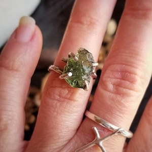 May include: A silver ring with a large, raw, green peridot gemstone. The ring is on a person's finger.