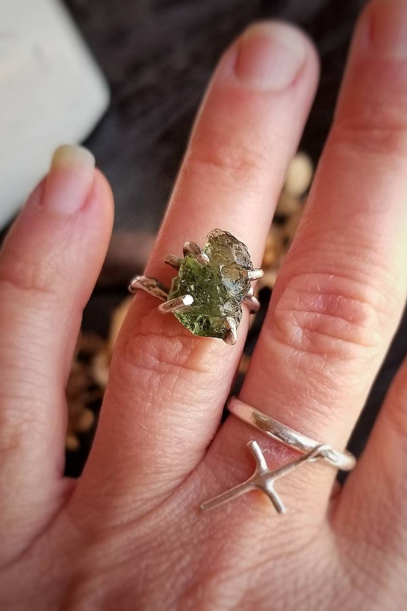 May include: A silver ring with a large, raw, green peridot gemstone. The ring is on a person's finger.
