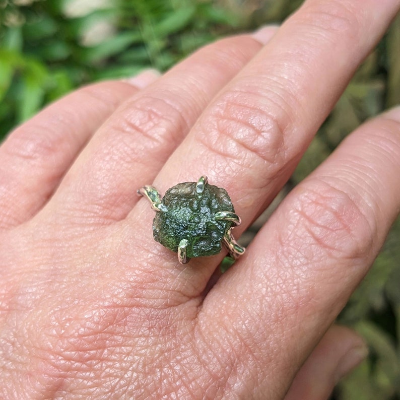 May include: A silver ring with a green, rough, and faceted gemstone. The ring is set in a simple, modern style.