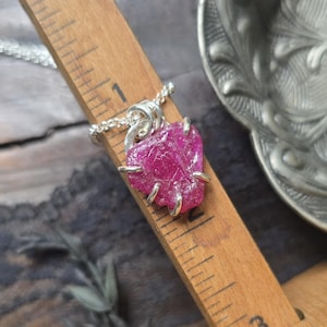 May include: A raw, deep pink gemstone pendant set in a silver-toned metal prong setting, suspended from a delicate silver chain. The pendant is positioned against a wooden ruler, showing a width of approximately 0.75 inches for the stone.