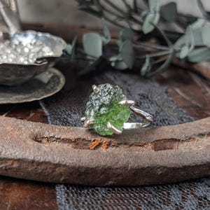 May include: A silver ring with a green, rough, natural gemstone. The ring is set on a rustic, brown surface.